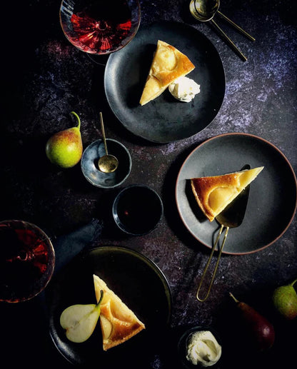 Dessert flat lay scene with slices of cheesecake, pears and red wine styled on a vinyl photography backdrop in mixed dark greys captured from real granite.