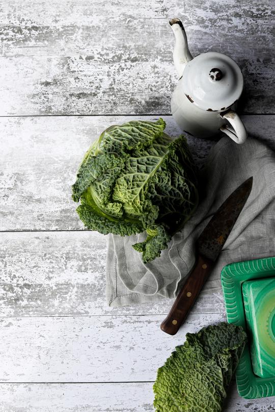 A flat lay of a white vintage teapot, fresh cabbages and an old knife styled on a wood effect vinyl photography backdrop with horizontal planks in warm grey and white.