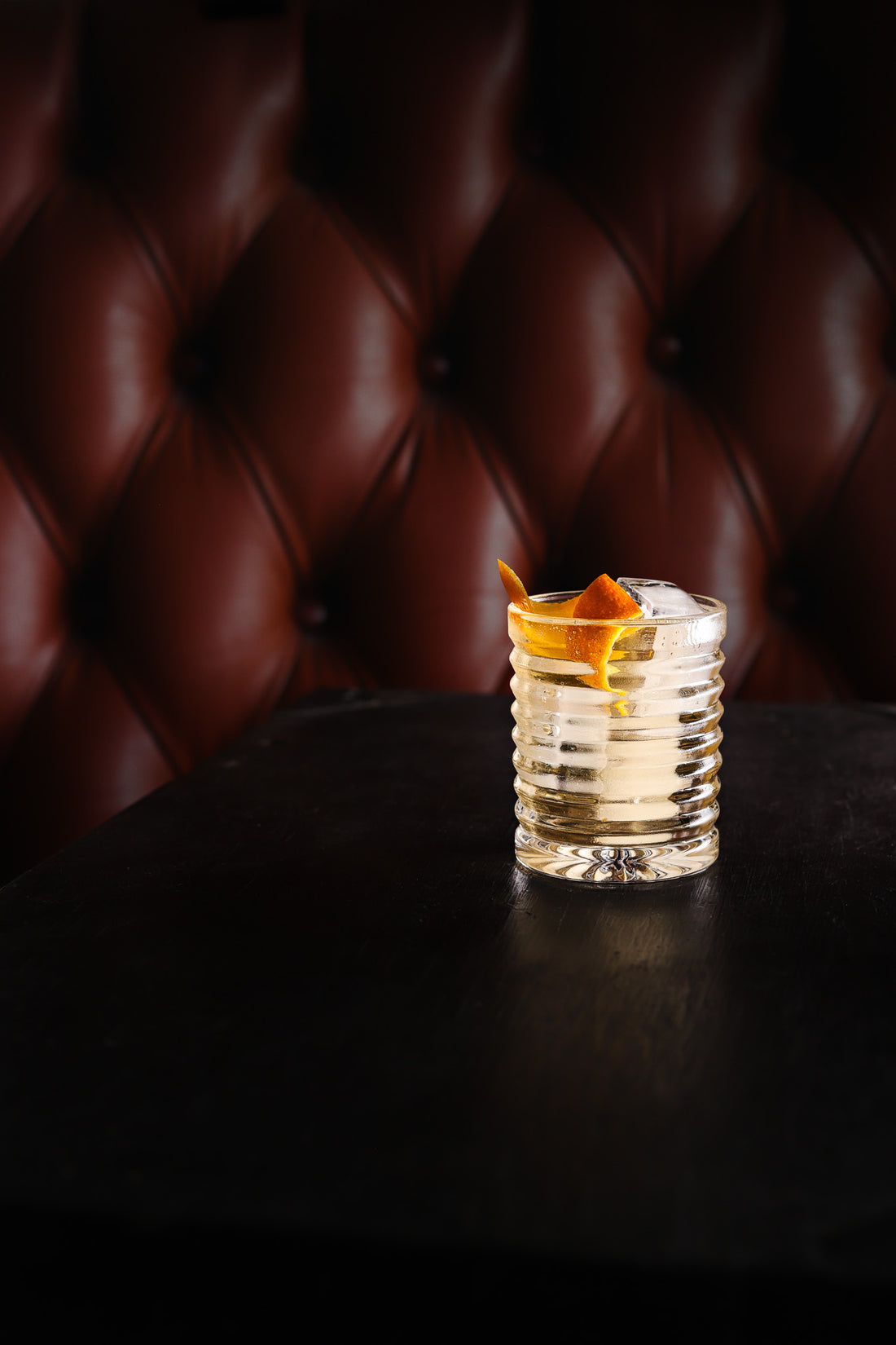 A whisky cockail in a ribbed tumbler with a slice of orange peel against a tan leather effect photography backdrop with button details, resembling a Chesterfield sofa.