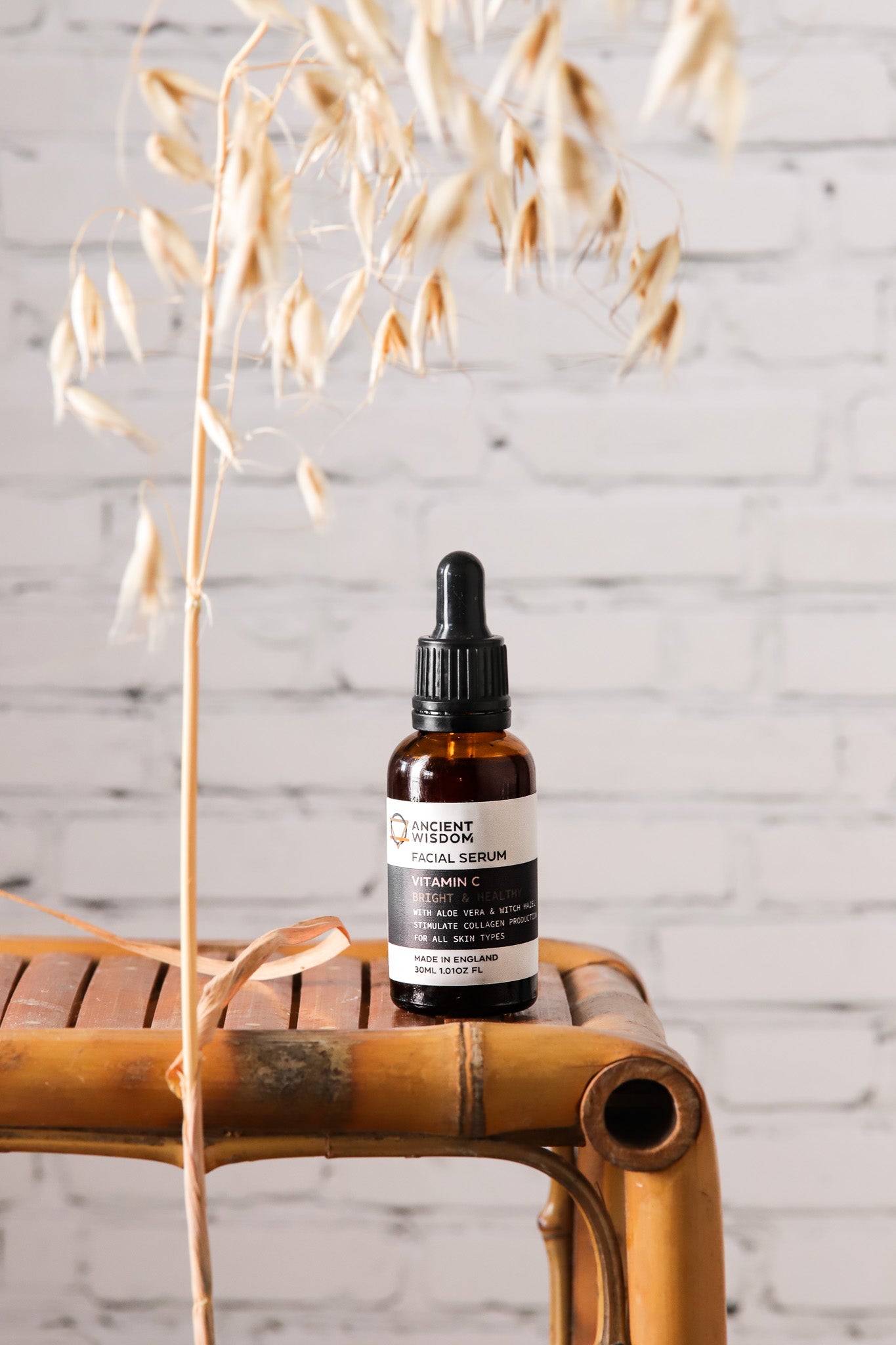 A facial serum against a white brick wall-effect vinyl photography backdrop with dried grasses in the foreground.
