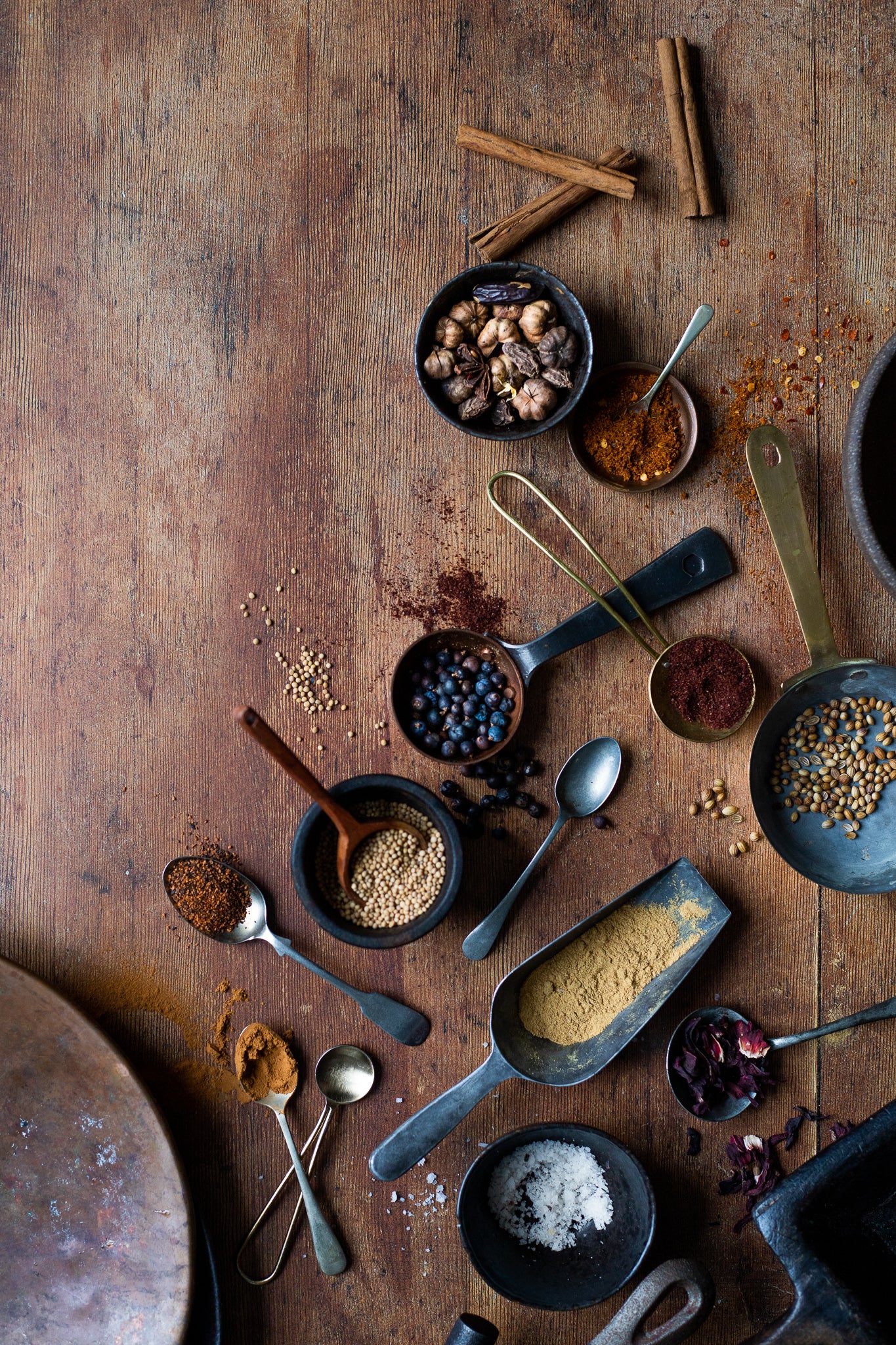 A styled image of a selection of vintage spoons filled with spices scattered across a wood-effect vinyl photography backdrop with natural color tones and visible grain details.