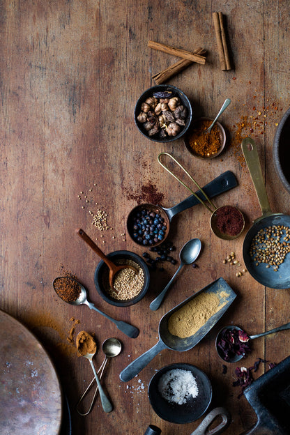 A styled image of a selection of vintage spoons filled with spices scattered across a wood-effect vinyl photography backdrop with natural color tones and visible grain details.