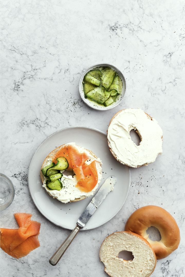 A white vinyl food photography backdrop with a subtle quartz or marble effect with plated salmon bagel flat lay.