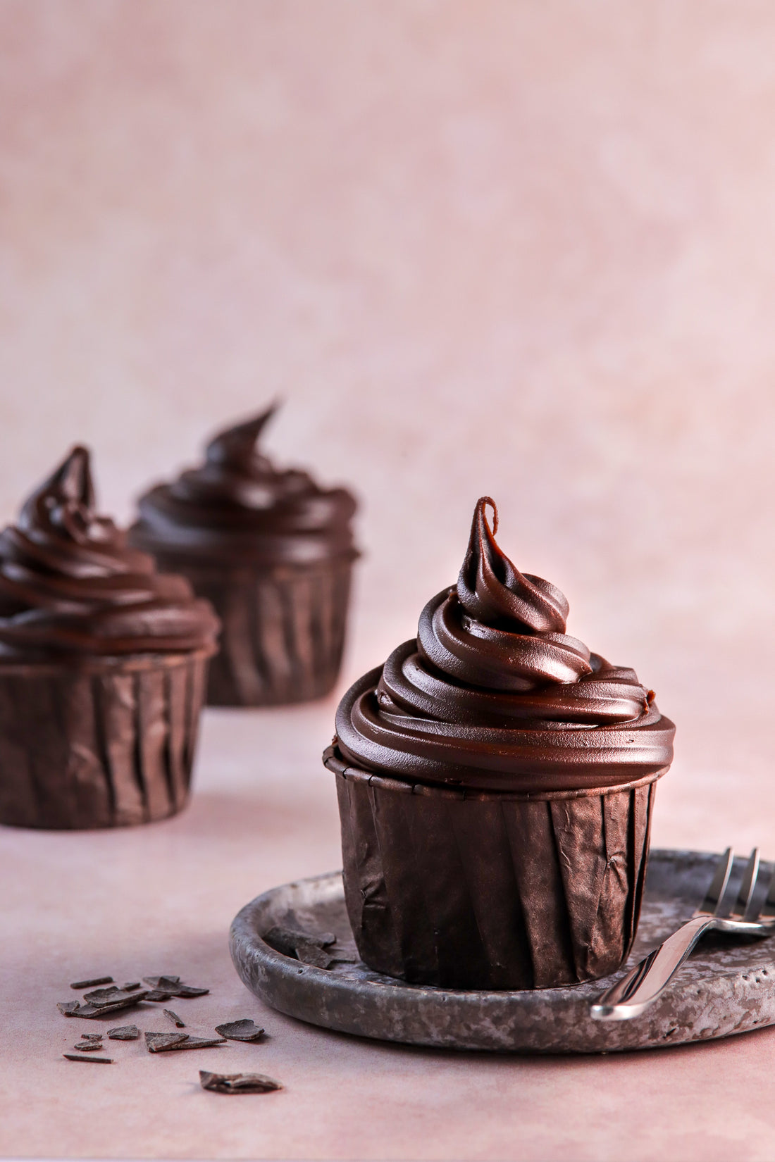 Image of three dark chocolate cupcakes on a blush pink marble effect photography backdrop.