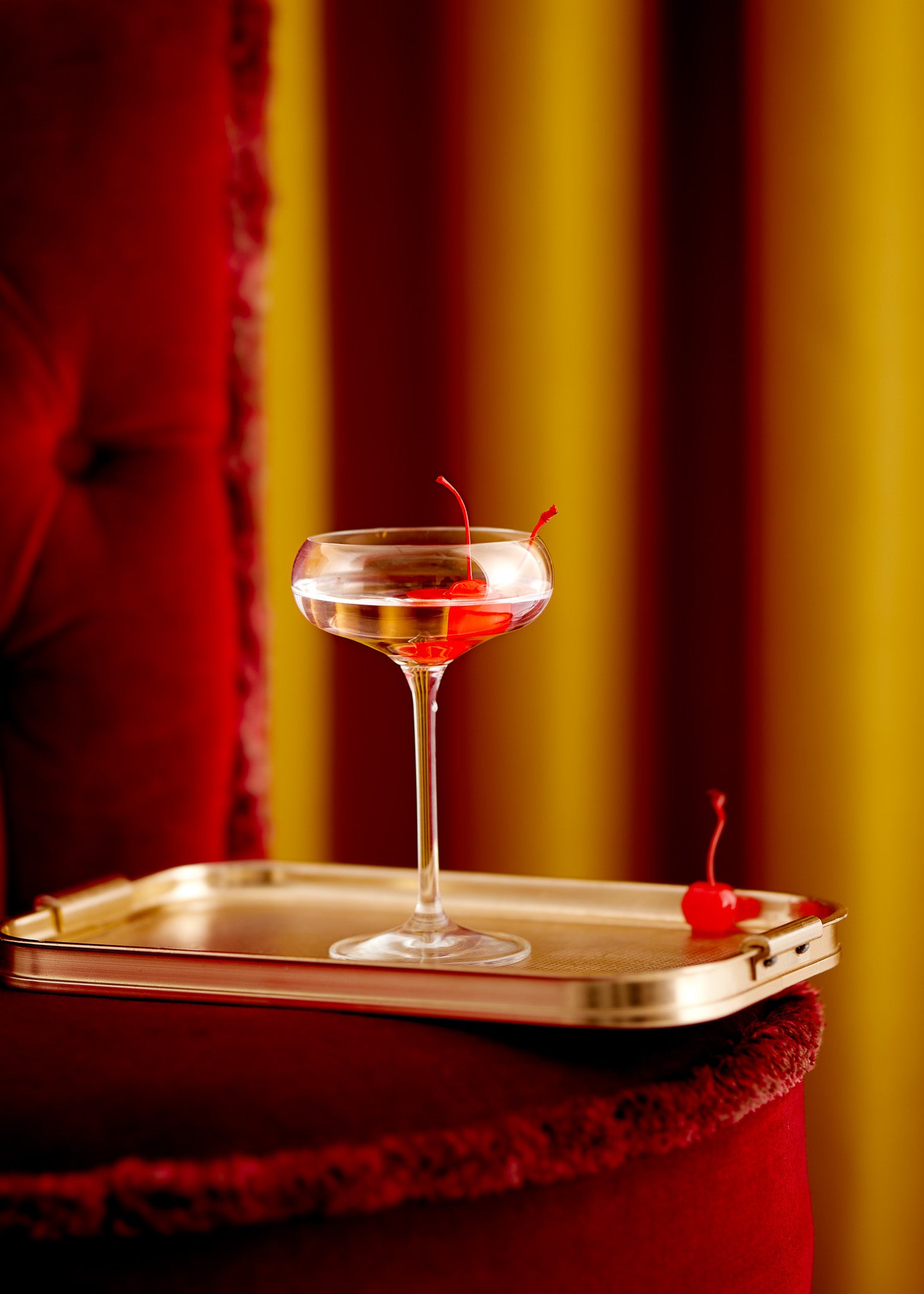 Cherry gin cocktail on a gold tray and red velvet sofa against a gold curtain-effect vinyl photography backdrop.