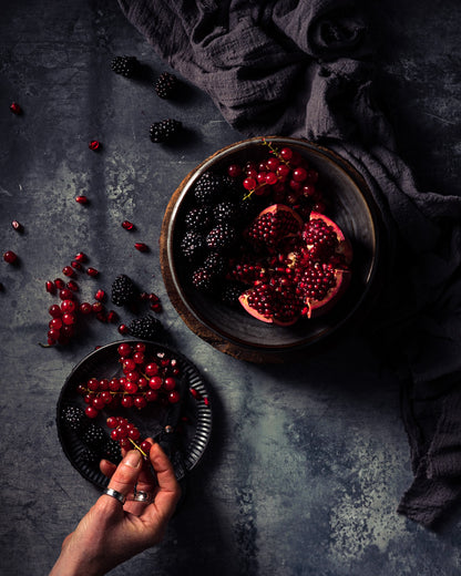Flay lay of forest fruit berries, blackberry pomegranate, styled on a grey vinyl photography backdrop with mottling and staining designed to resemble an aged zinc table surface, originally designed for cookbook cover photography.