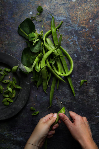 A flat lay image of hands cutting fresh green beans styled on a brown textured slate-effect vinyl photography backdrop.
