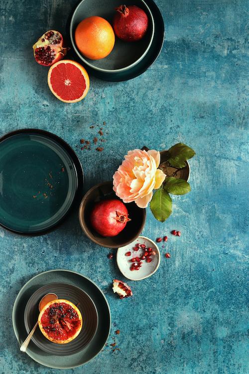 A textured photography backdrop with a mottled effect featuring shades of turquoise, blue, and teal with grey flecks with blue ceramic plates, fruit and flower flat lay.