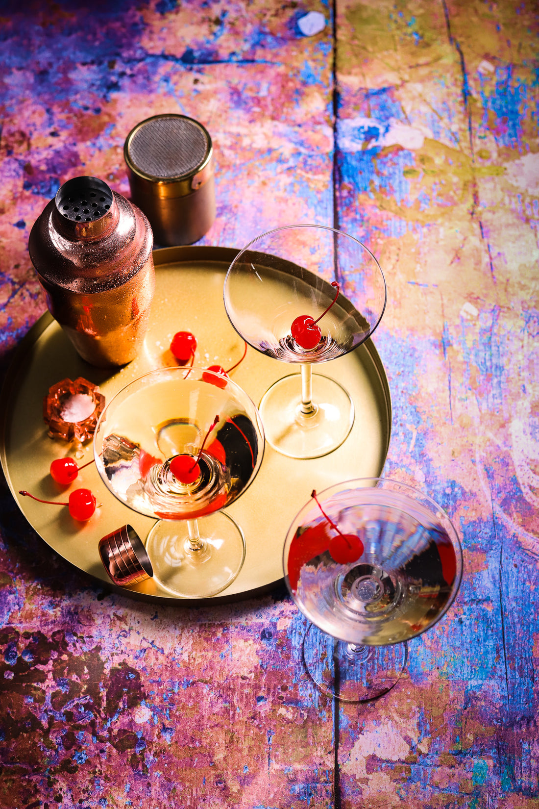 Gold cocktail tray and mixers with coupe glasses shot on a neon paint effect vinyl photography background featuring a mix of orange, yellow, blue, purple, and pink.