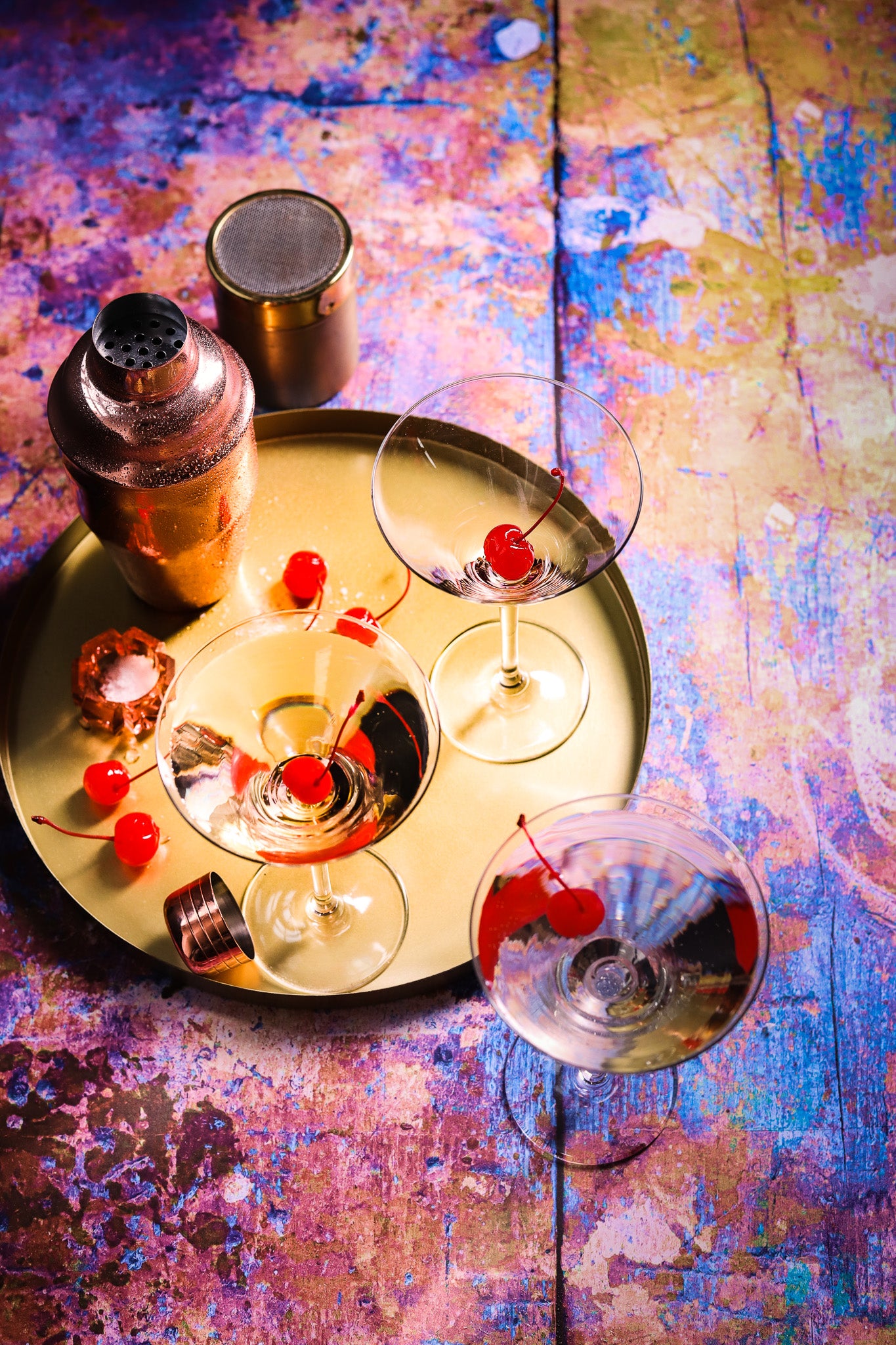 Gold cocktail tray and mixers with coupe glasses shot on a neon paint effect vinyl photography background featuring a mix of orange, yellow, blue, purple, and pink.