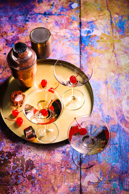 Gold cocktail tray and mixers with coupe glasses shot on a neon paint effect vinyl photography background featuring a mix of orange, yellow, blue, purple, and pink.