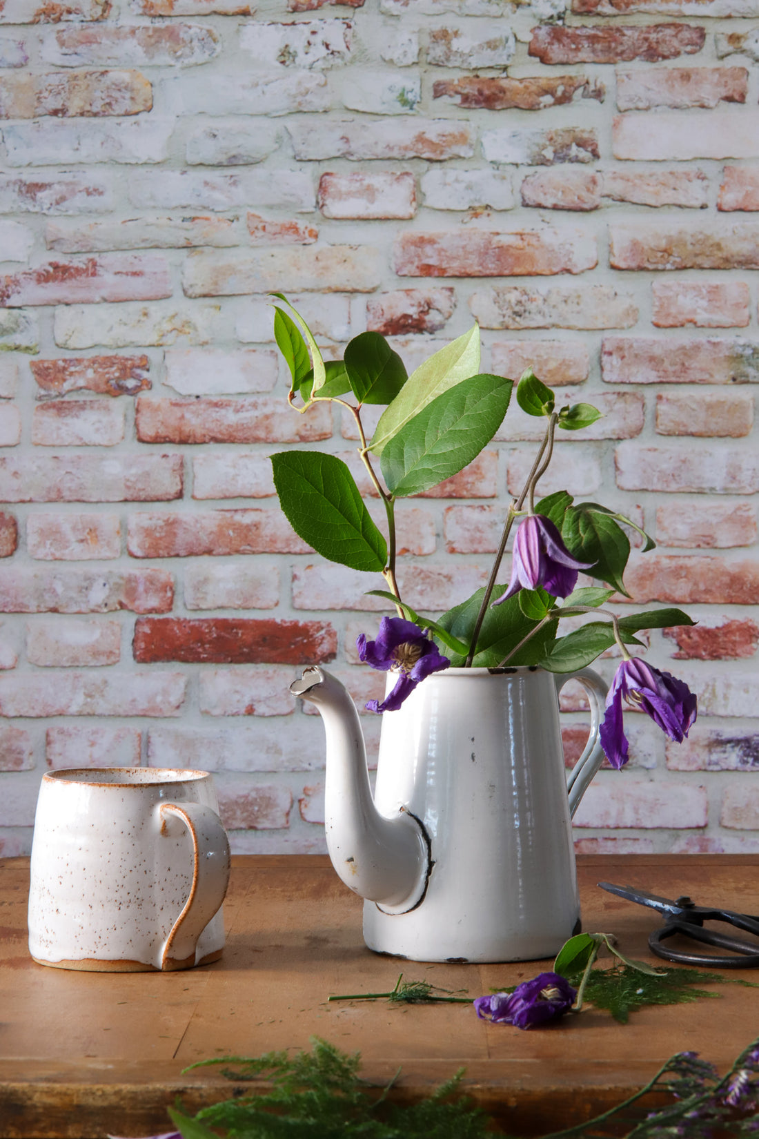 Vintage jug with purple flowers styled against a photography backdrop in a mix of red, grey, and white colors, simulating a rough brick wall texture.