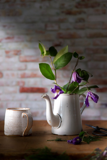 Vintage mug and a jug with wilted purple flowers styled against a photography backdrop in a mix of red, grey, and white colors, simulating a rough brick wall texture.