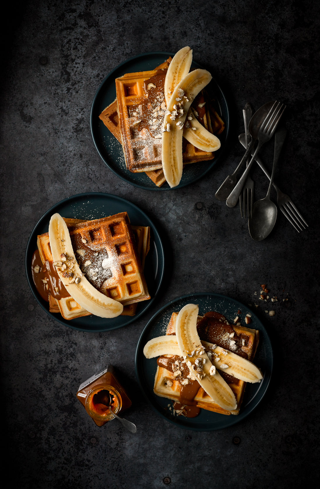 A flat lay of banana waffles styled on a dark grey metal patina effect photography backdrop with a textured surface featuring original circular rivets.