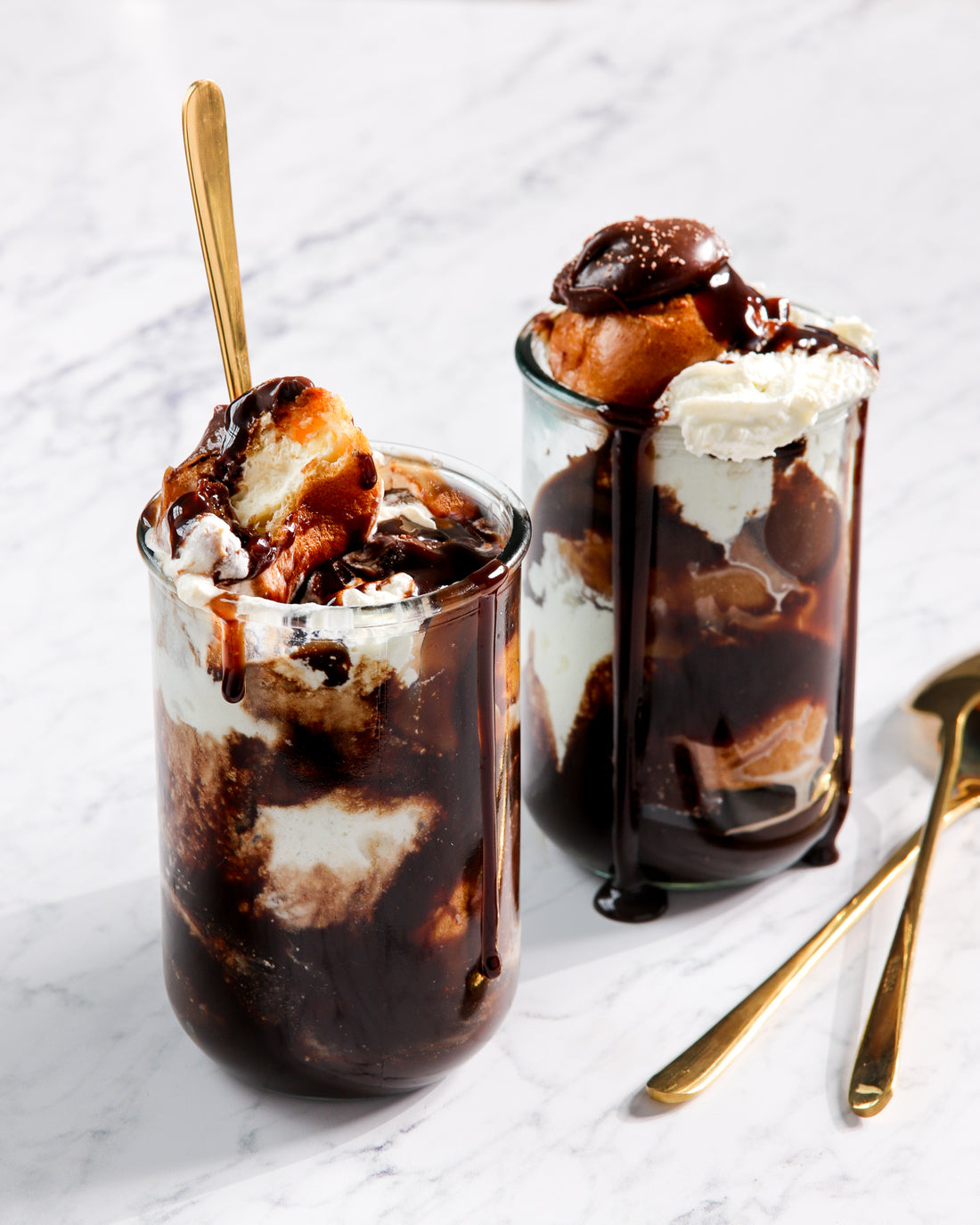 Two glasses of chocolate icecream sundaes styled on a grey and white marble effect vinyl photography backdrop.