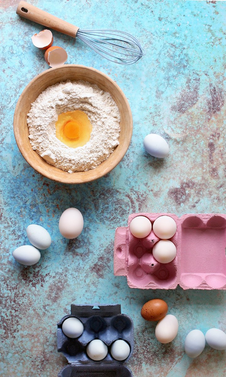 Overhead view of a bowl of flour with a cracked egg, surrounded by a whisk, eggshells, loose eggs, and colourful egg cartons shot against a copper patina effect vinyl photography backdrop in shades of turquoise and rust.