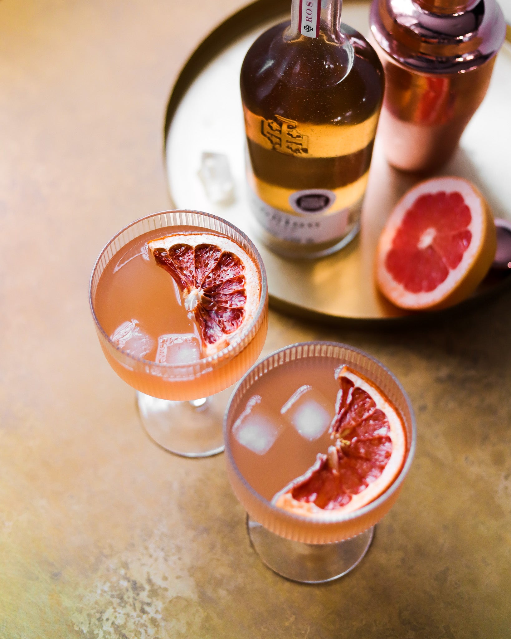 A tray of whisky and grapefruit cocktails styled on a golden brass metal effect photography backdrop with a softly mottled texture.