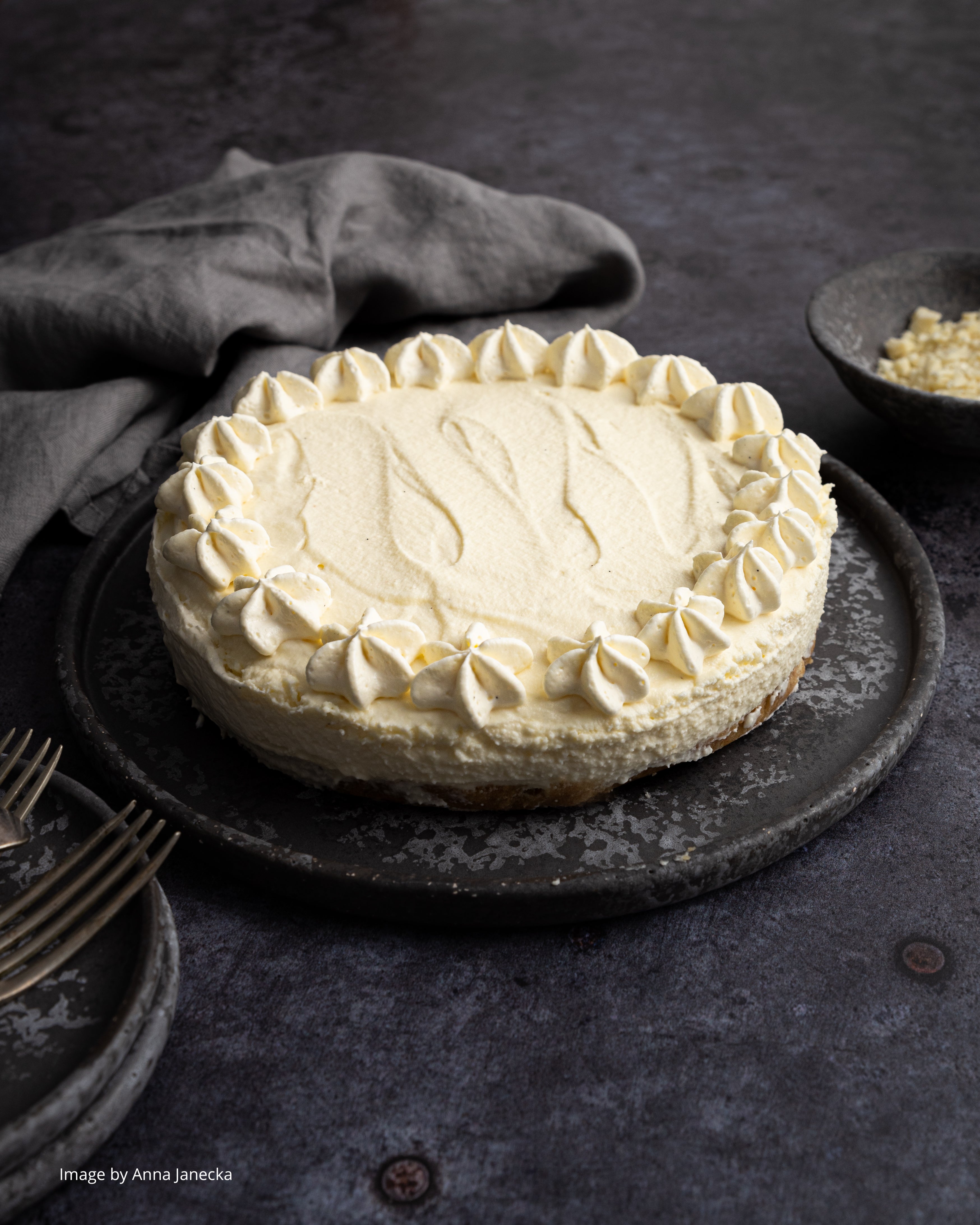 A white handmade cheesecake styled on a dark grey metal patina effect photography backdrop with a textured surface featuring original circular rivets.